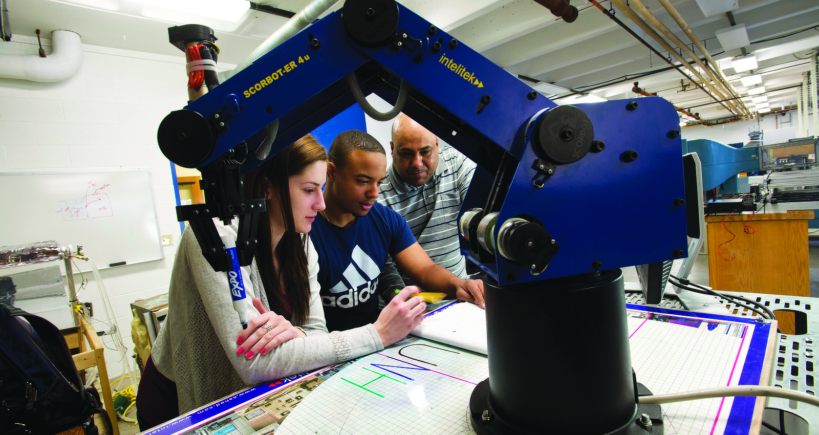 A female and male student collaborating over technical plans while operating a jointed mechanical robotic arm in an engineering laboratory.