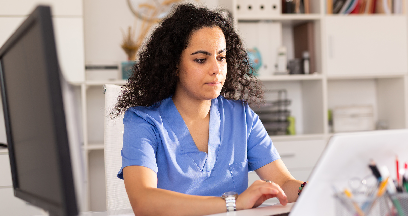 A female healthcare professional wearing blue scrubs typing on a laptop in an office setting.