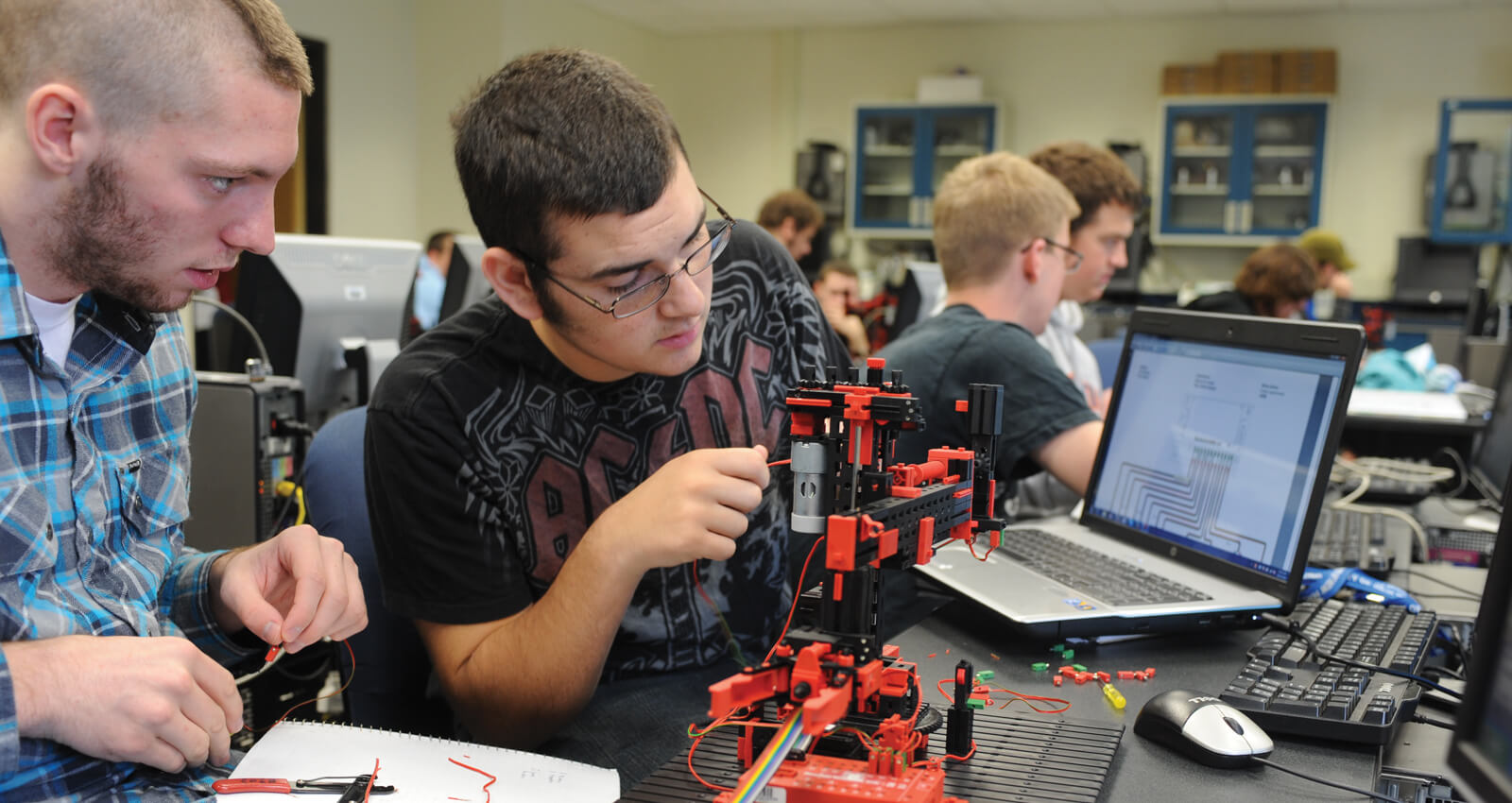 A male student adjusting wiring on a red modular robotic mechanism inside an engineering laboratory.