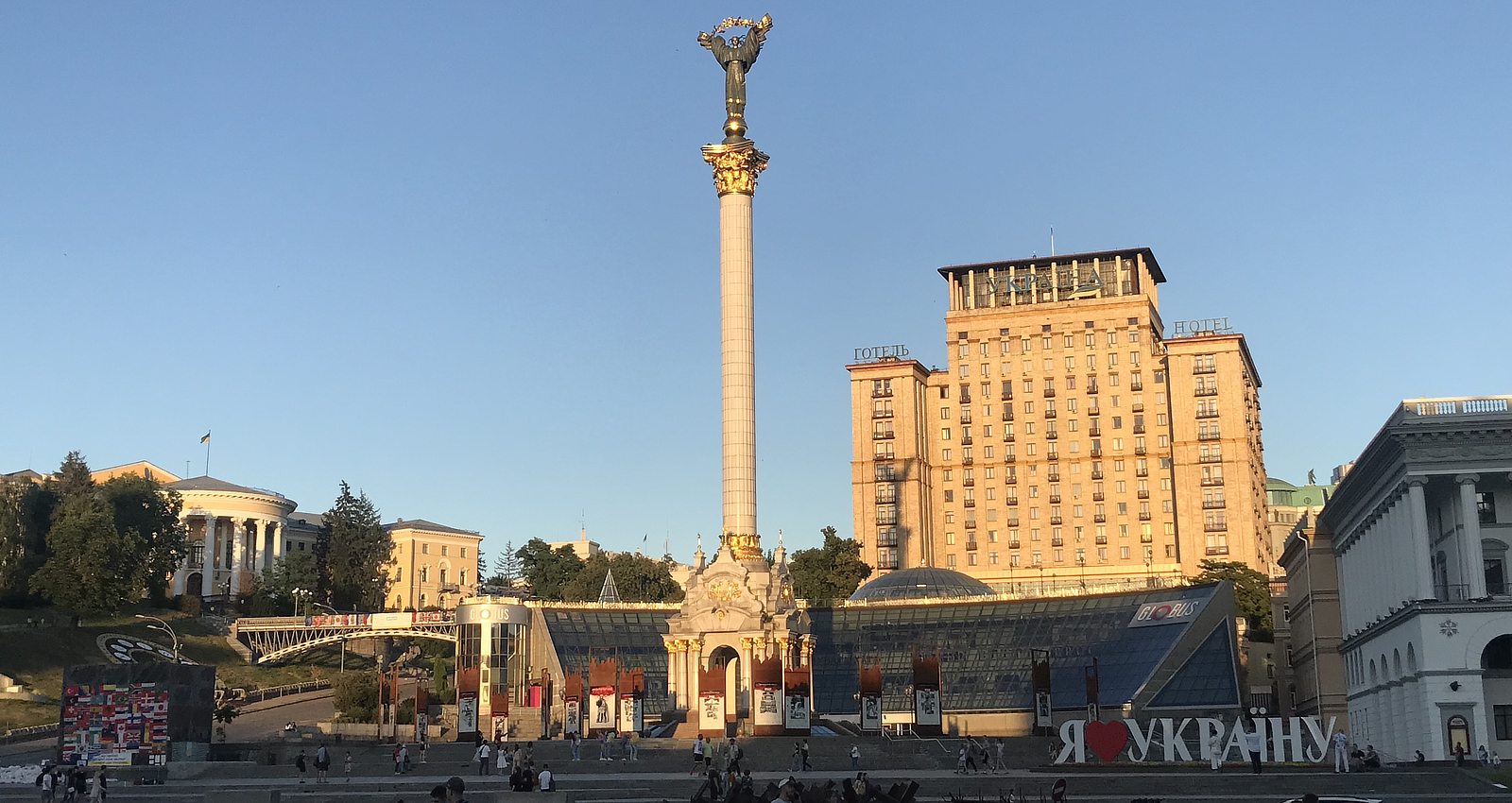 A wide shot of Independence Square in Kyiv, Ukraine, featuring the Independence Monument tall column, the Hotel Ukraine building, and decorative signage reading 'I Love Ukraine' in Ukrainian script.