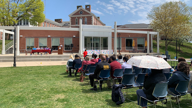 A crowd gathers to listen to a speaker outside.