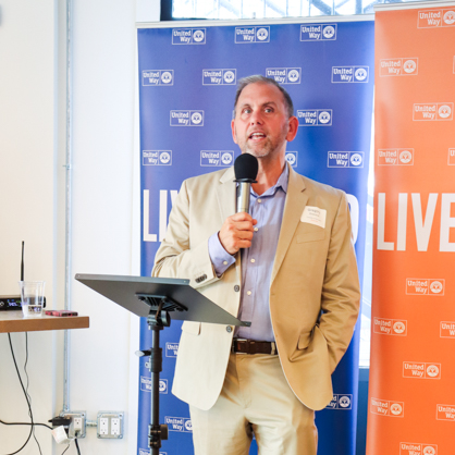 Greg Overend accepted the /U+0026#x27;Live United/U+0026#x27; award on behalf of the University of New Haven. He holds a microphone and stands in front of a red and blue step and repeat