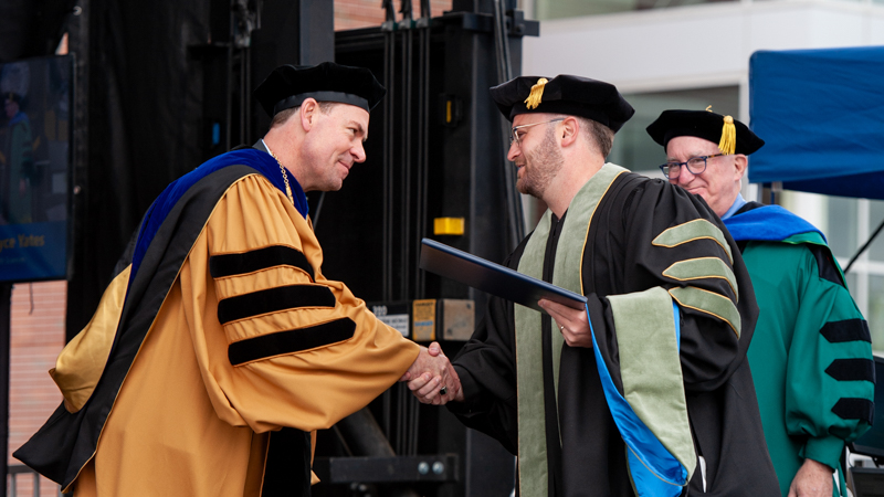 Brian A. Marks, J.D., Ph.D., looks on as his son, Aaron S. Marks ’24 DHSc shakes hands with President Jens Frederiksen, Ph.D., at Commencement